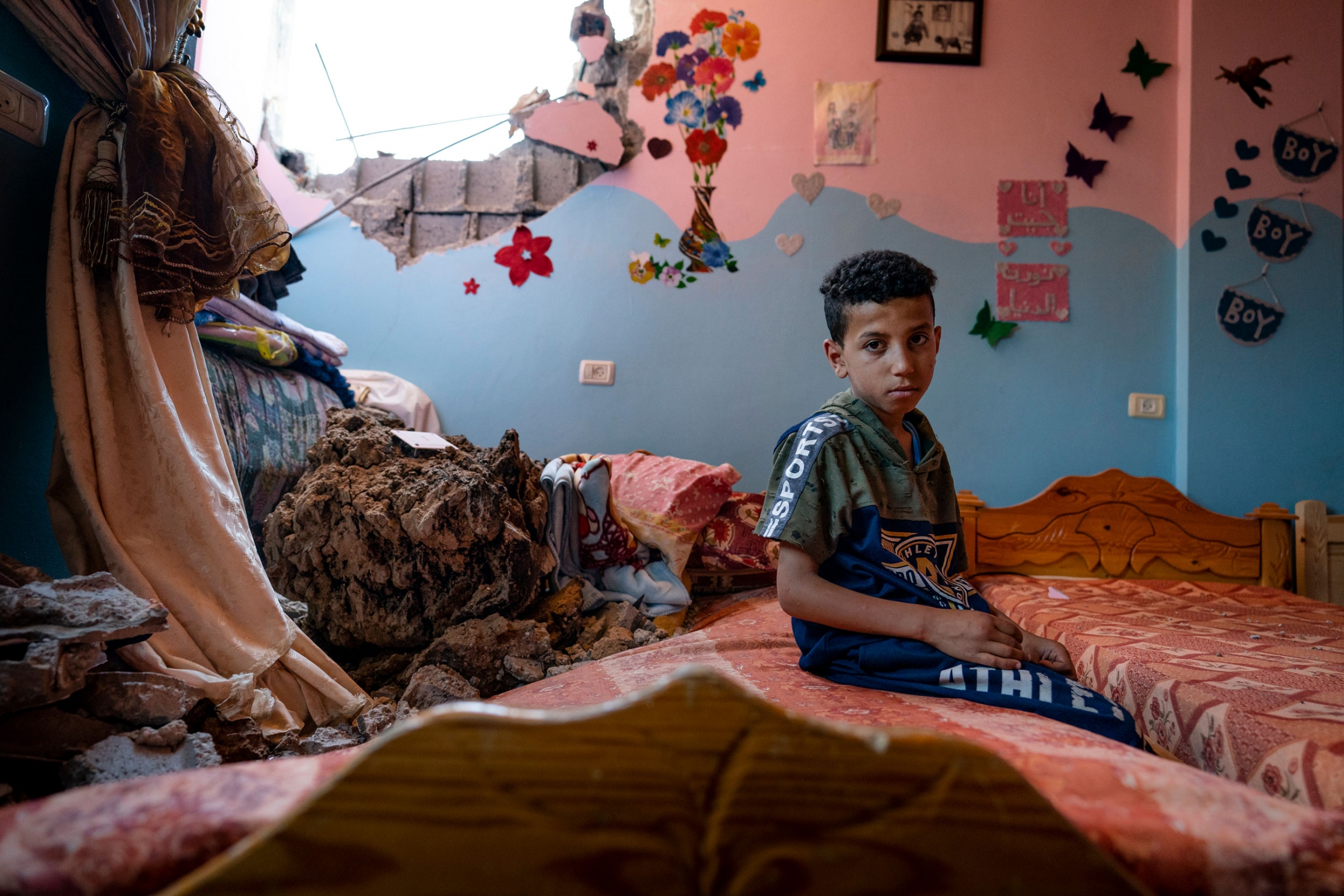 Ibrahim Al-Masri, 10, sits for a portrait in his bedroom that was damaged when an airstrike destroyed the neighboring building in Beit Hanoun, Gaza Strip, May 26, 2021. A young boy sits on a bed in a room with pink and blue painted walls and a large hole in the wall.
