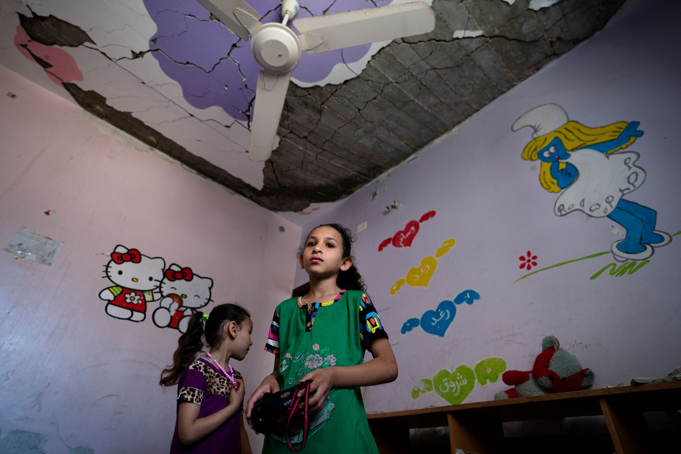 Suroq Al-Masri, 9, and her sister Razan, 4, stand for a portrait in their bedroom that was severely damaged when an airstrike destroyed a neighboring building, in Beit Hanoun, Gaza Strip, May 26, 2021. Two young girls are shown in a room with a smurf cartoon on the wall and a damaged ceiling.