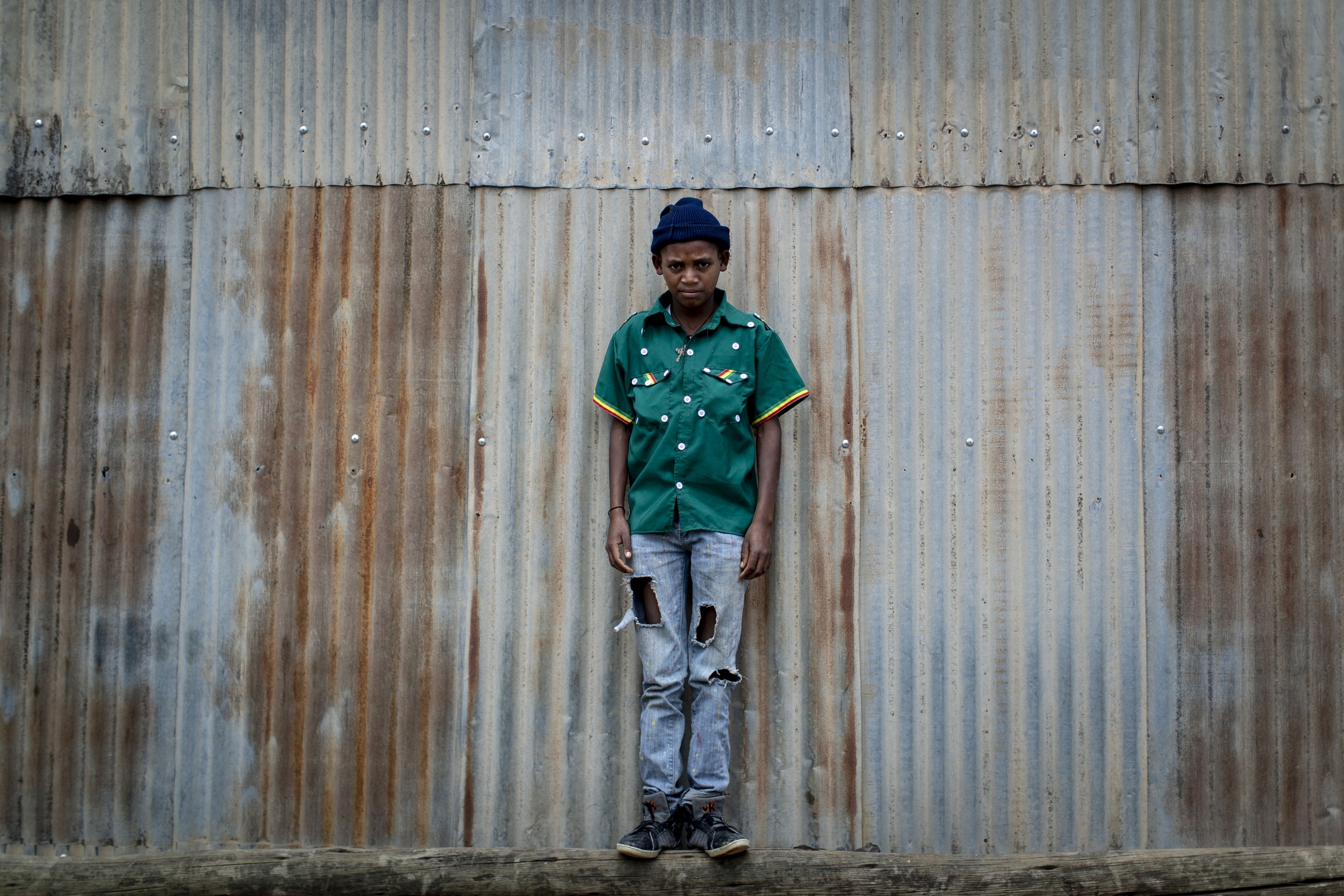 Gebremedhin Gebreslassie, 12, who fled from fighting in the town of Hawzen, stands next to a metal shack at a reception center for the internally displaced in Mekele, in the Tigray region of northern Ethiopia, on Sunday, May 9, 2021. boy stands against corrugated steel wearing a green cultural shirt with buttons and jeans.