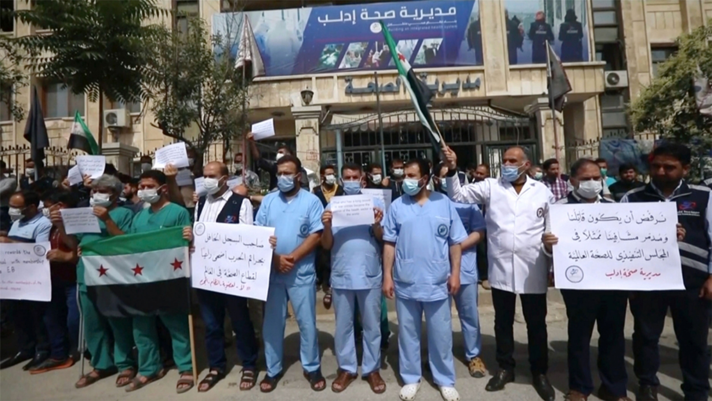 In this image taken from video, dozens of medical workers protest a decision to grant Syrian President Bashar al-Assad’s government a seat on the executive board of the World Health Organization, in Idlib, Syria Doctors stand in front of a hospital holding signs