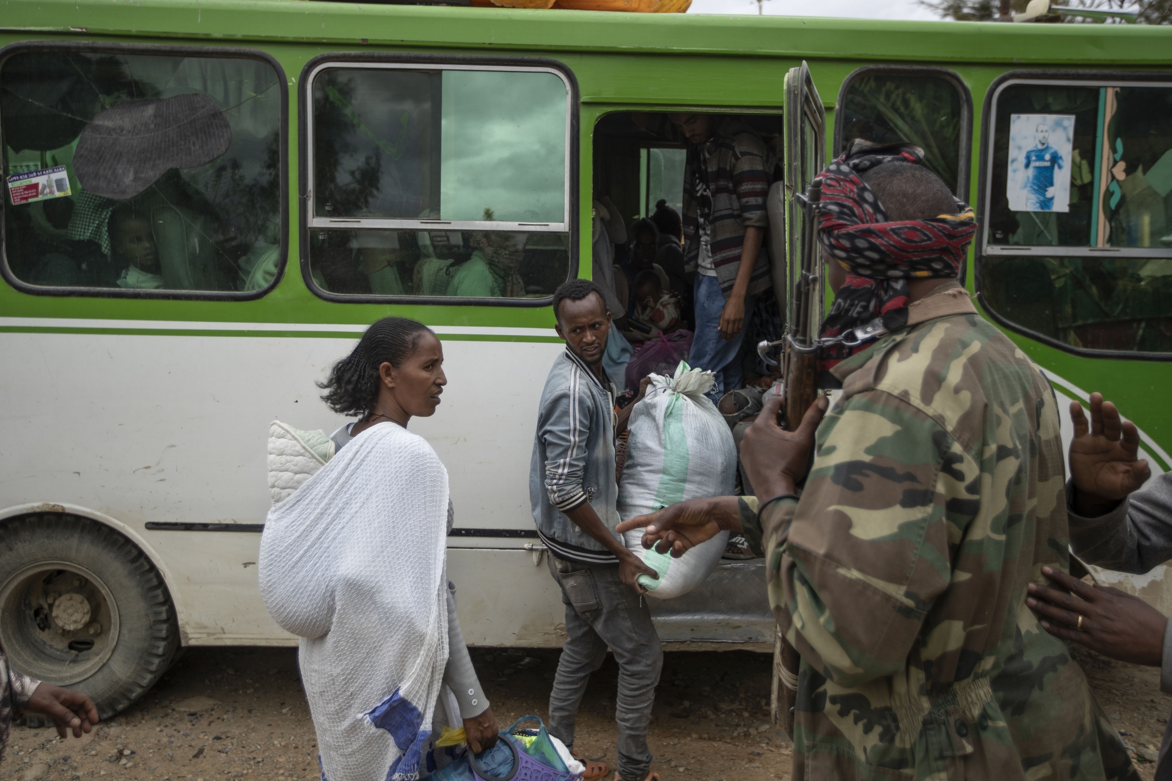 People prepare to depart on a bus as a fighter loyal to the Tigray People's Liberation Front (TPLF), right, stands guard in the town of Hawzen, then-controlled by the group, in the Tigray region of northern Ethiopia, on Friday, May 7, 2021. Travelers get on a bus.