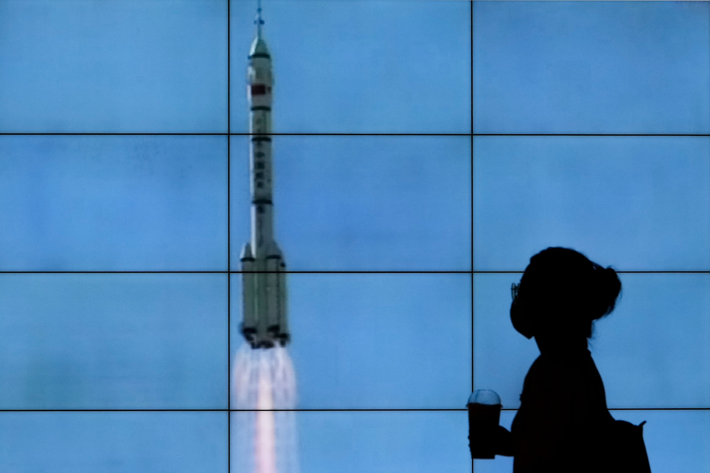 A woman wearing a face mask is silhouetted as she walks by a TV screen showing CCTV live telecast of the Long March-2F Y12 rocket carrying a crew of Chinese astronauts, at a shopping mall in Beijing. A woman is shown in shadow standing in front of a screen showing the Chinese spacehip lifting off.
