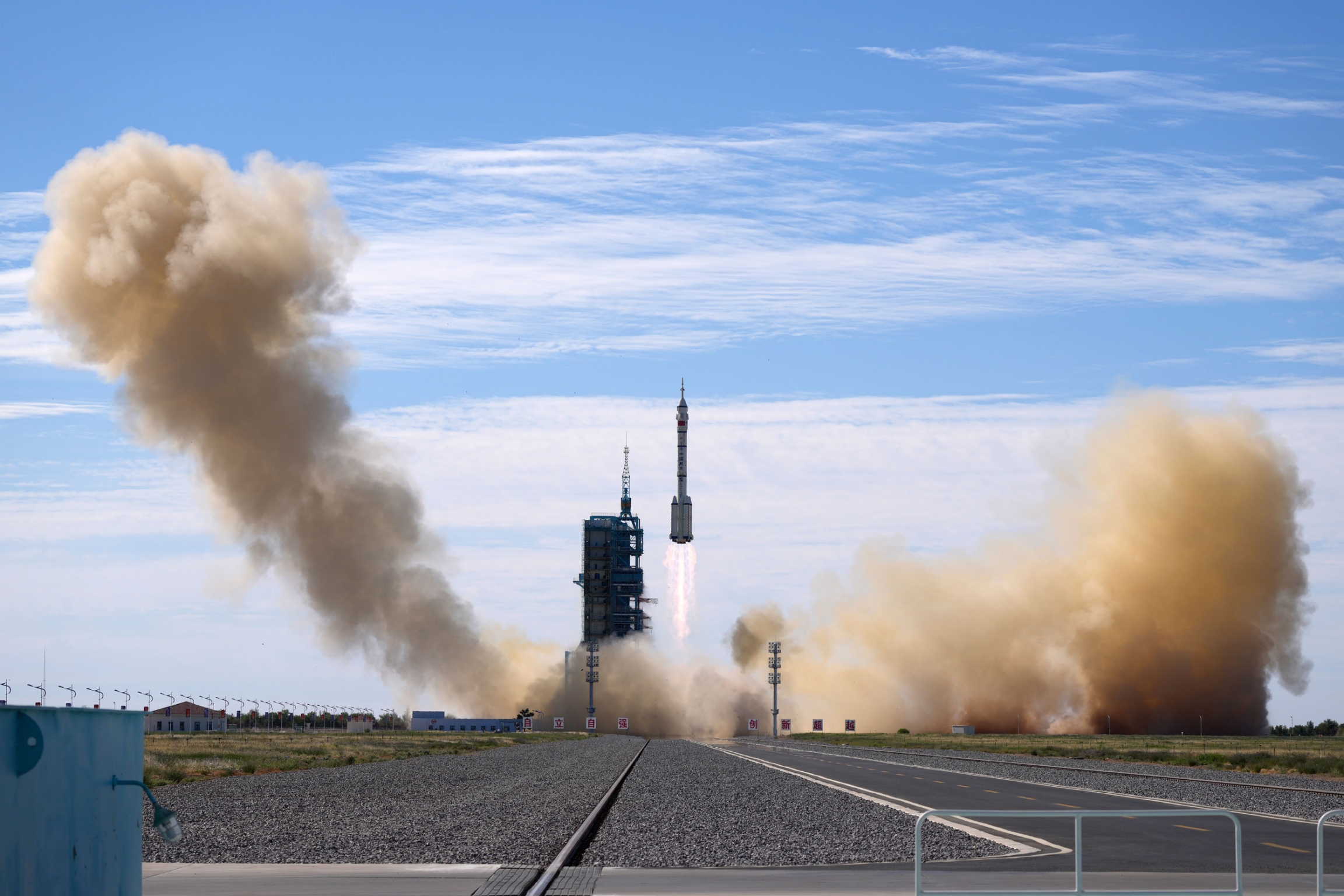 A Long March-2F Y12 rocket lifts off at the Jiuquan Satellite Launch Center. A spaceship is shown in the far distance being lifted into the sky via rockets with smoke billowing.