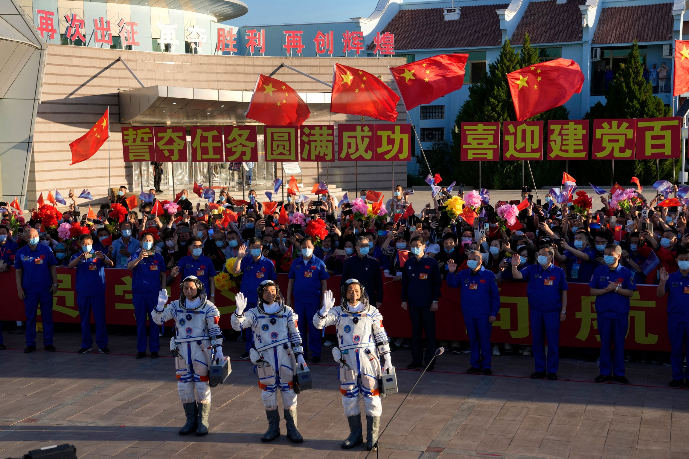 Chinese astronauts, from left, Tang Hongbo, Liu Boming, and Nie Haisheng wave at the Jiuquan Satellite Launch Center in Jiuquan in northwestern China. Three Chinese astronauts are shown waving with a large crowd of people waving the Chinese flag behind them.