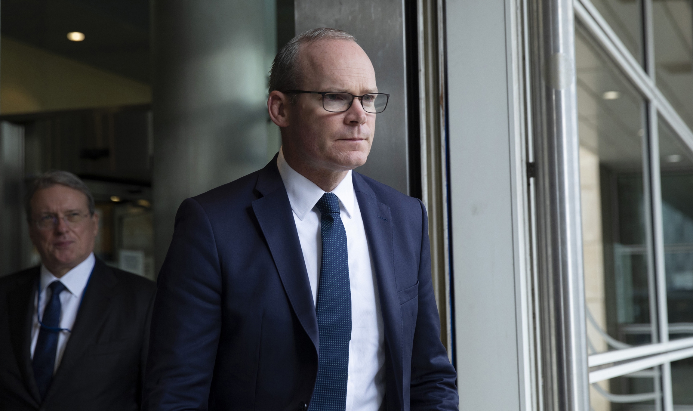 In this Friday, Sept. 27, 2019 file photo, Irish Foreign Minister Simon Coveney leaves EU headquarters in Brussels. A white man wears a blue suit and glasses.