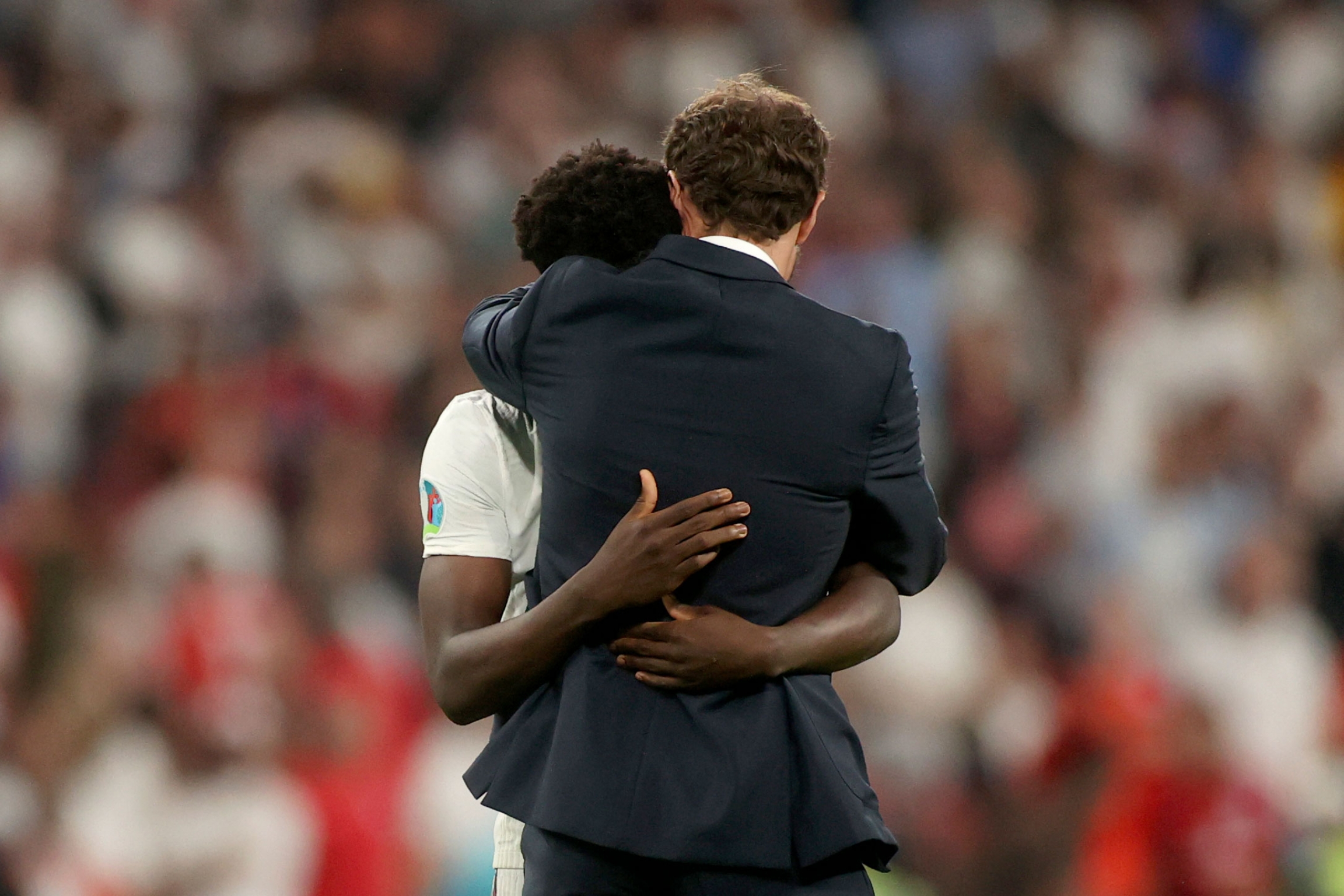 England's manager Gareth Southgate embraces Bukayo Saka after he failed to score a penalty during a penalty shootout. England's manager Gareth Southgate is shown hugging Bukayo Saka on the soccer pitch with fans shown in blurred focus in the distance.