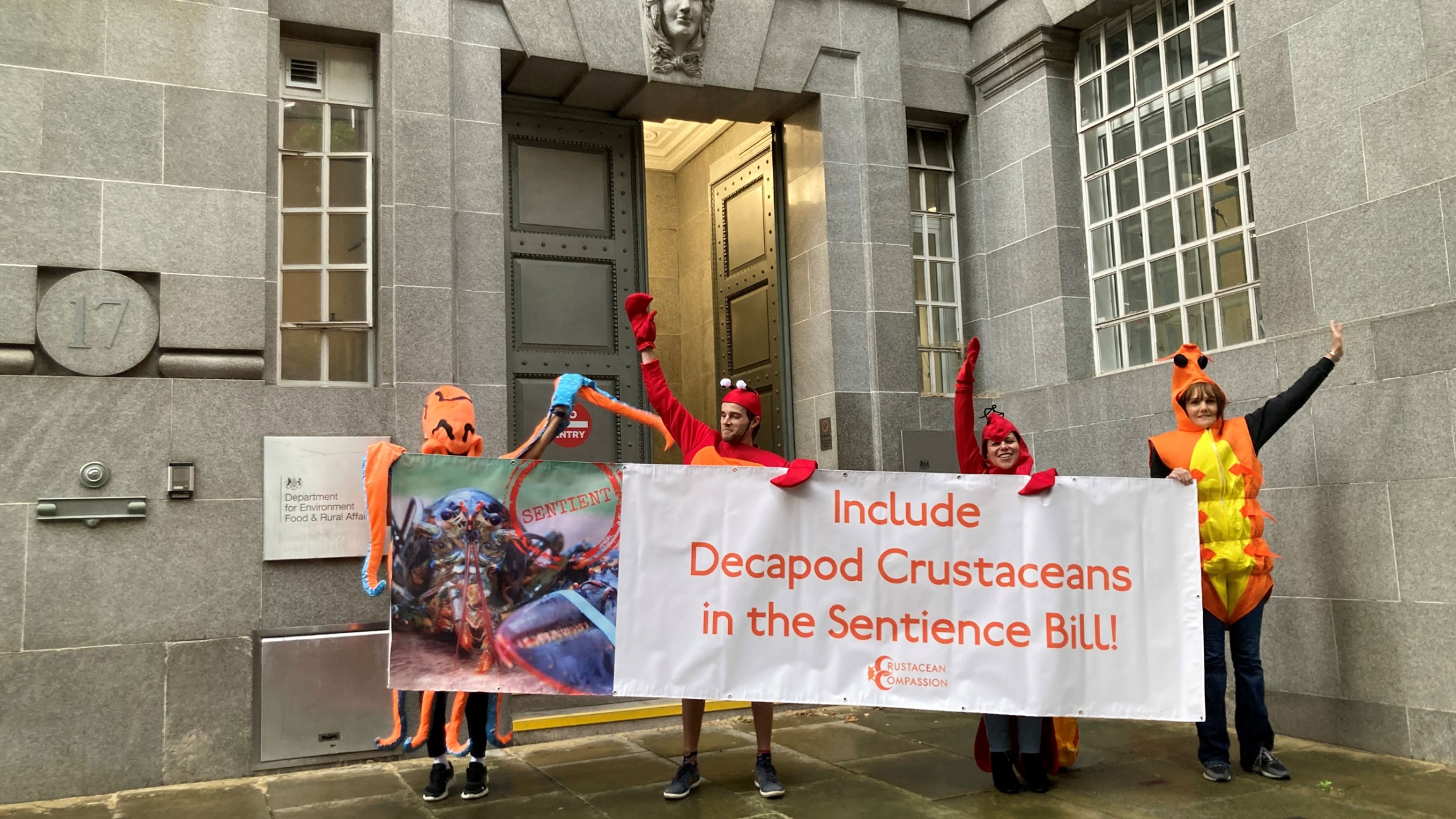 Crustacean Compassion activists cheer with a banner advocating for the sentience of shellfish. Activists dressed in shellfish costumes cheer with a banner.