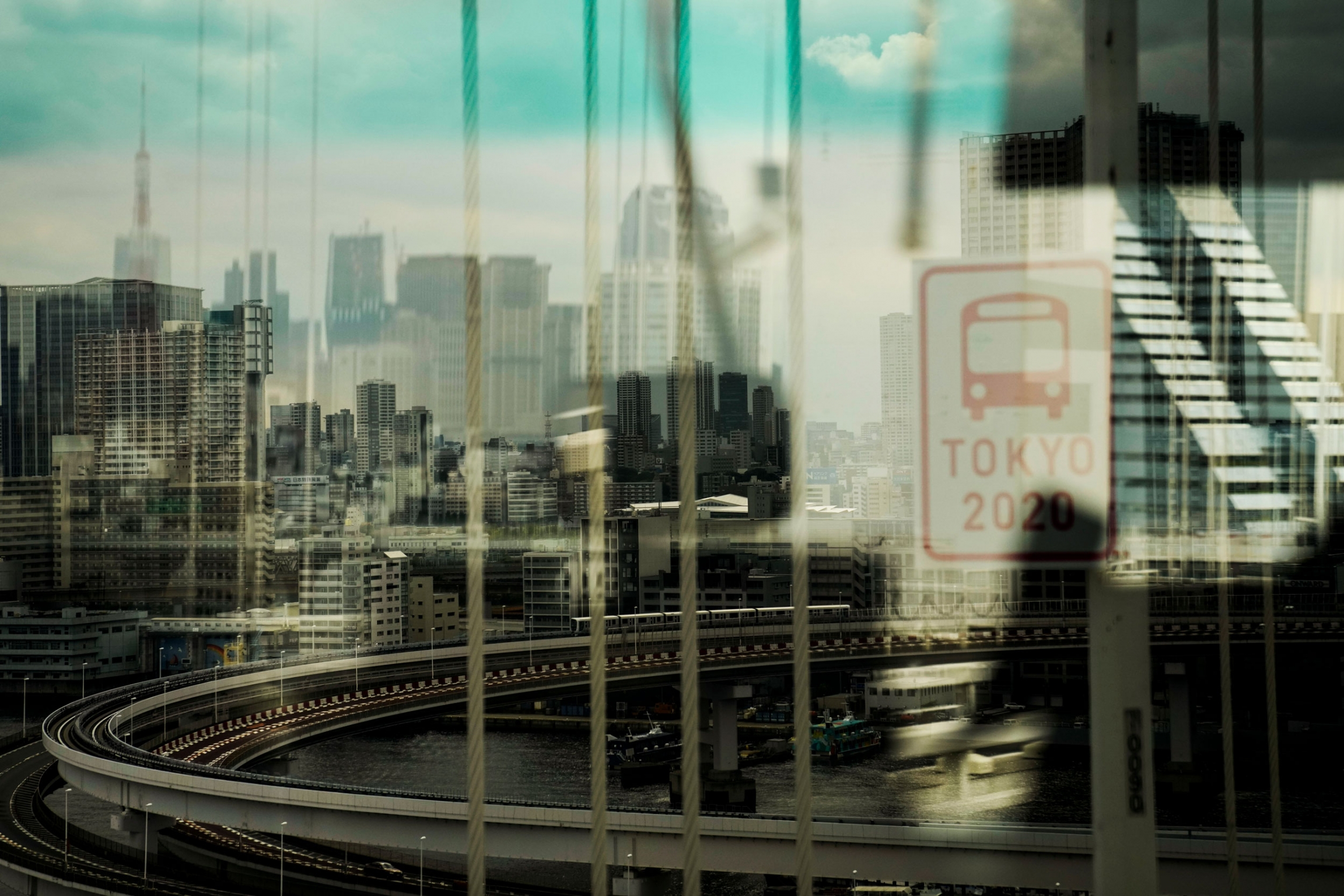 The skyline of Tokyo is seen through a bus window from the Rainbow Bridge ahead of the 2020 Summer Olympics. A major roadway and elevated train track along with the Tokyo skyline are shown blurred slightly via a bus window.