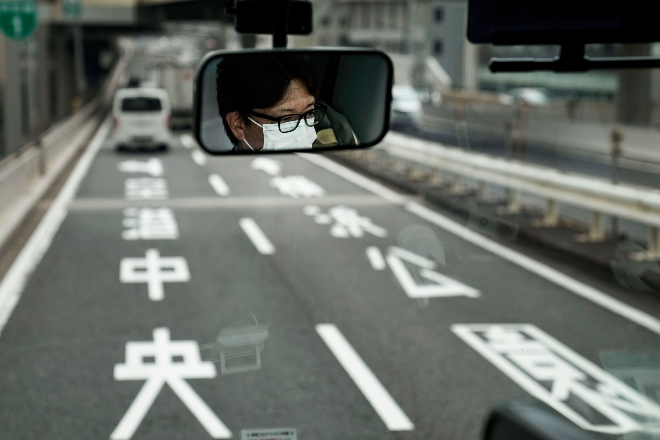A bus driver is reflected in the mirror in Tokyo. The face of a bus driver is shown covered in a white mask with a two lane road ahead.
