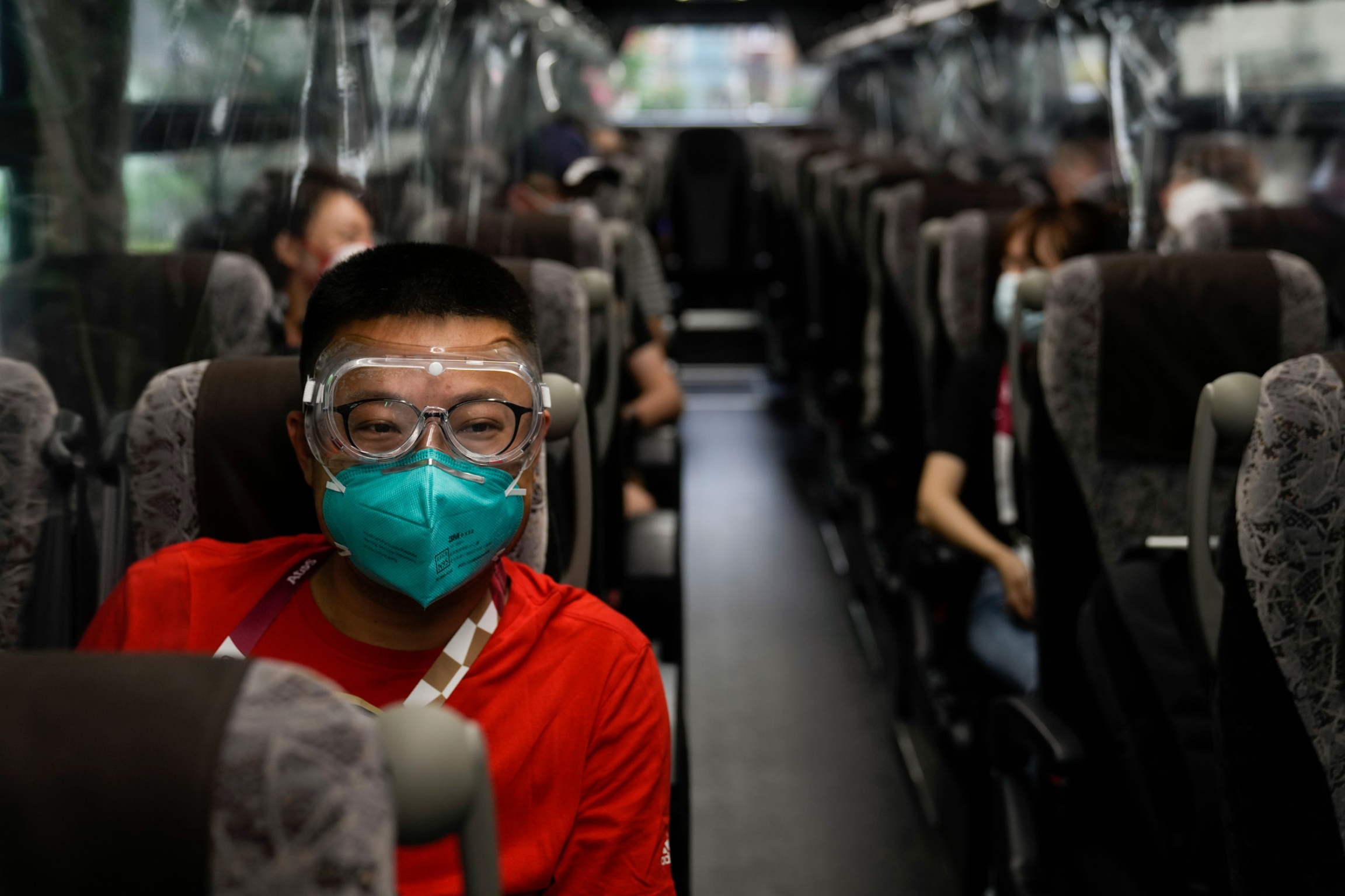 Hua Zhang, of the China Media Group, wears protective goggles while riding a media bus ahead the 2020 Summer Olympics. A man is shown wearing a red shirt, face mask and clear goggles while riding a bus.