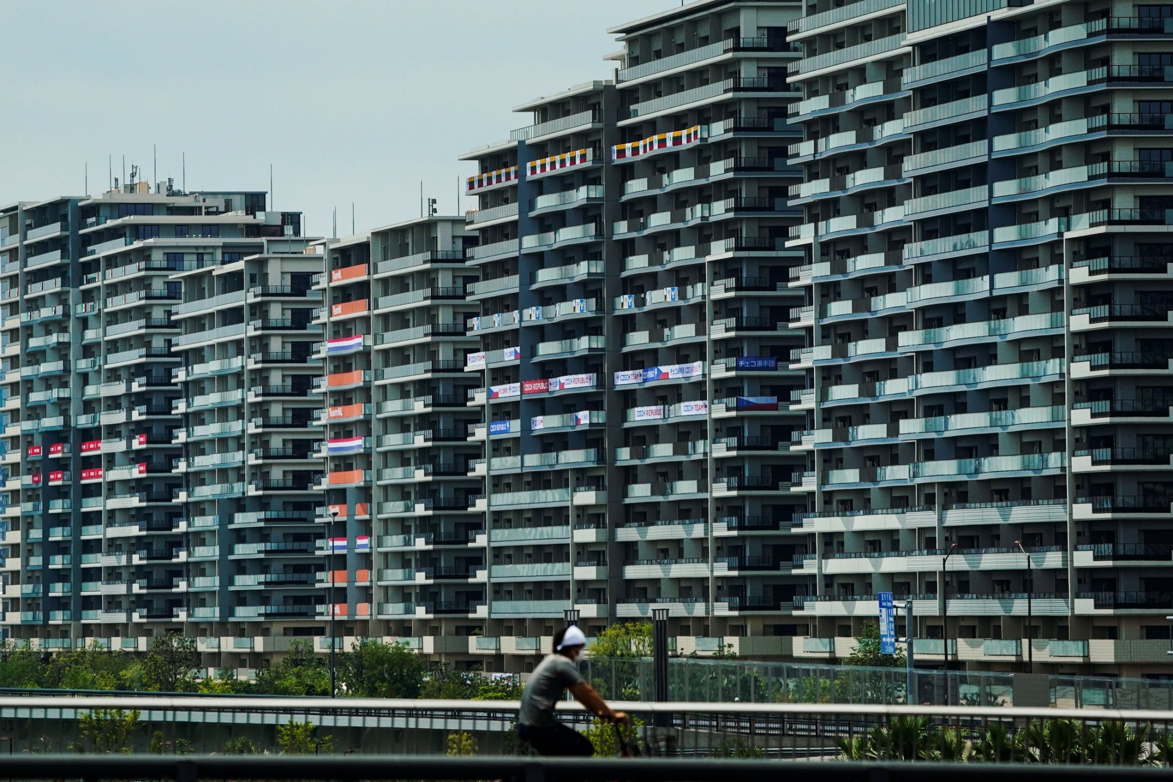A man rides his bike near the athlete's village for the 2020 Summer Olympics and Paralympics in Tokyo. A row of tall buildings are shown, many with national flags hanging from the balconies.