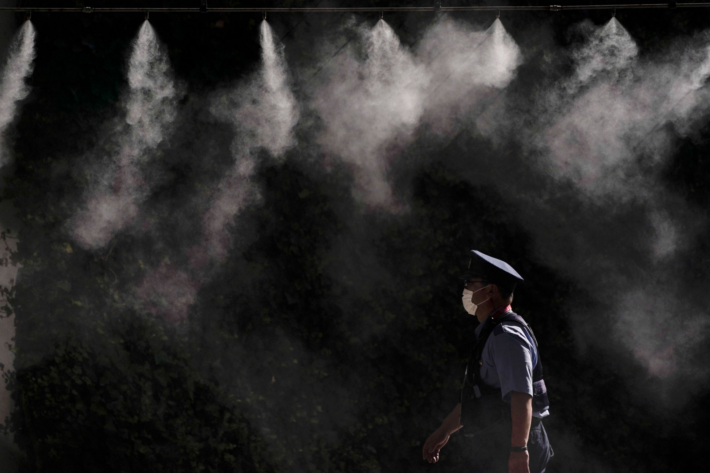 A cooling system sprays a mist over a police officer outside the main press center of the 2020 Summer Olympics. A police officer is shown wearing a white mask and walking under several misters spraying water.