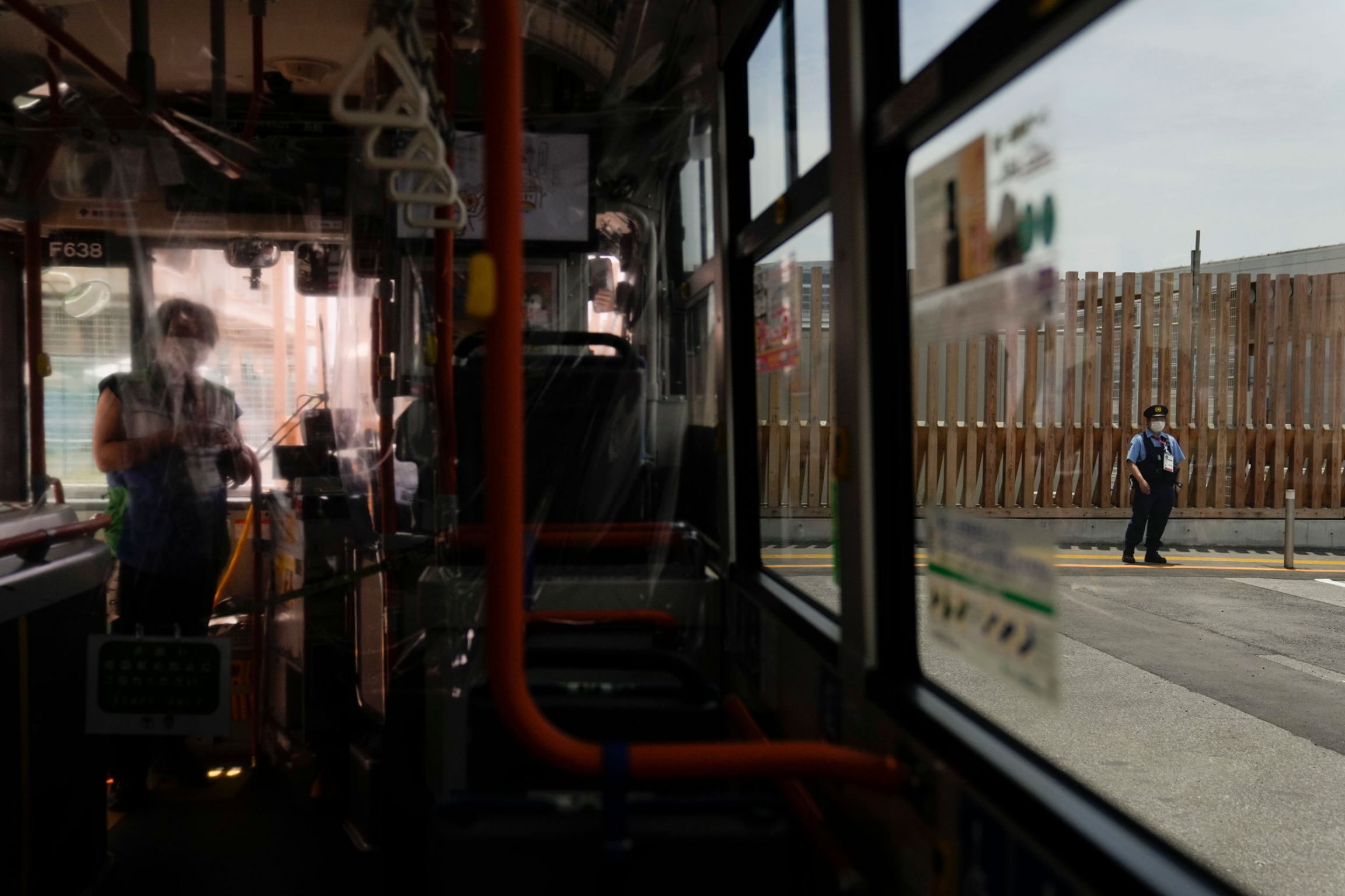 A police officer, right, patrols near the entrance to the athlete's village for the 2020 Summer Olympics and Paralympics. A police office is shown through the window of a bus standing guard in front of a metal barrier.
