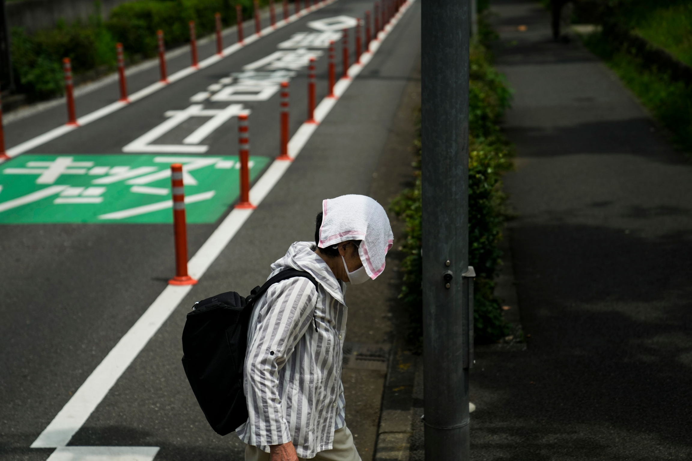 A woman walks with a small towel over her head to shield from the sun ahead the 2020 Summer Olympics. A woman is shown with a towel over her head and covering her face with a roadway in the distance.