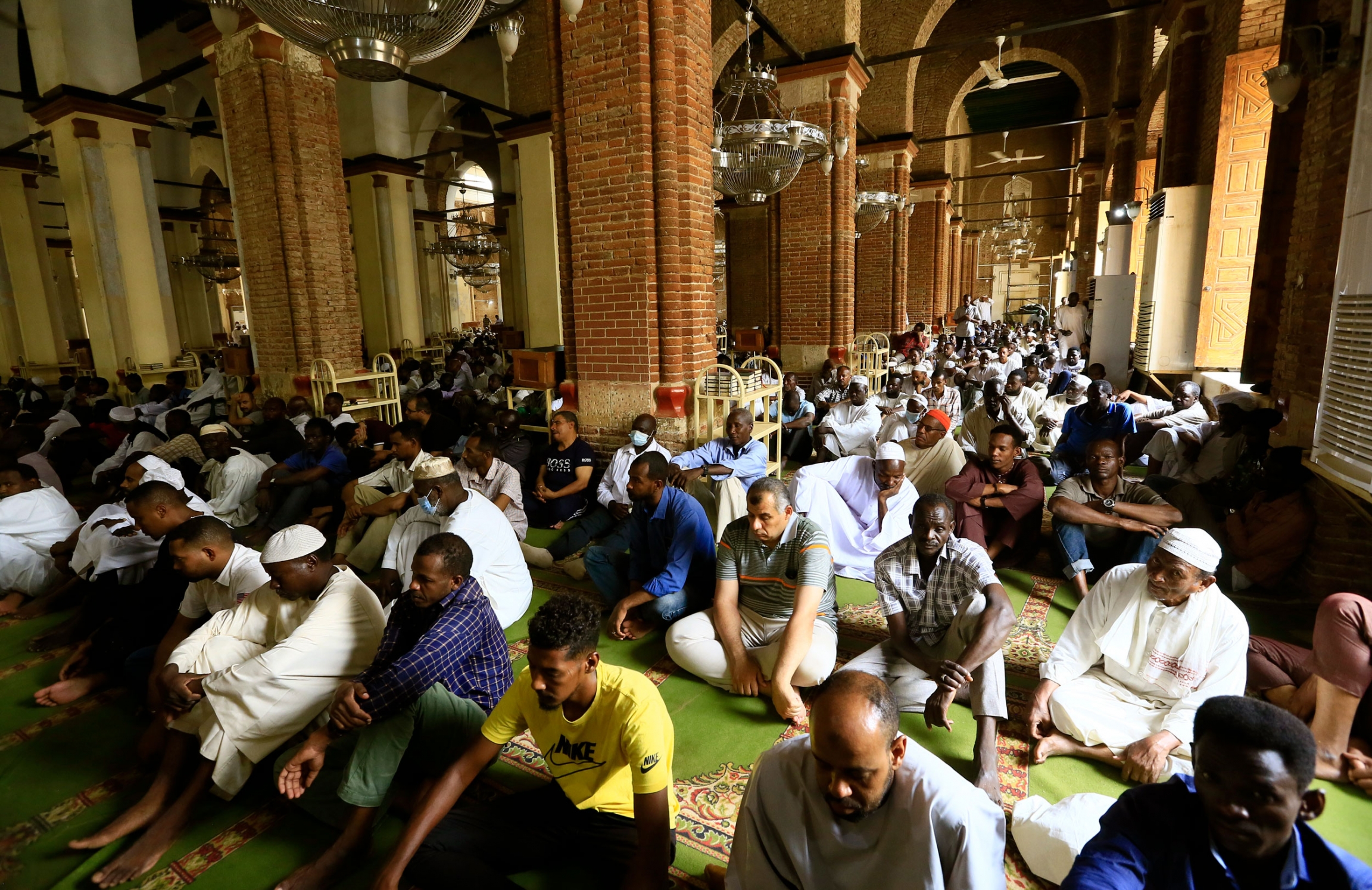 People gathered for Friday prayers at the Grand Mosque in Khartoum, Sudan. A large group of people are show sitting on the floor of the Grand Mosque with large brick columns down the middle.