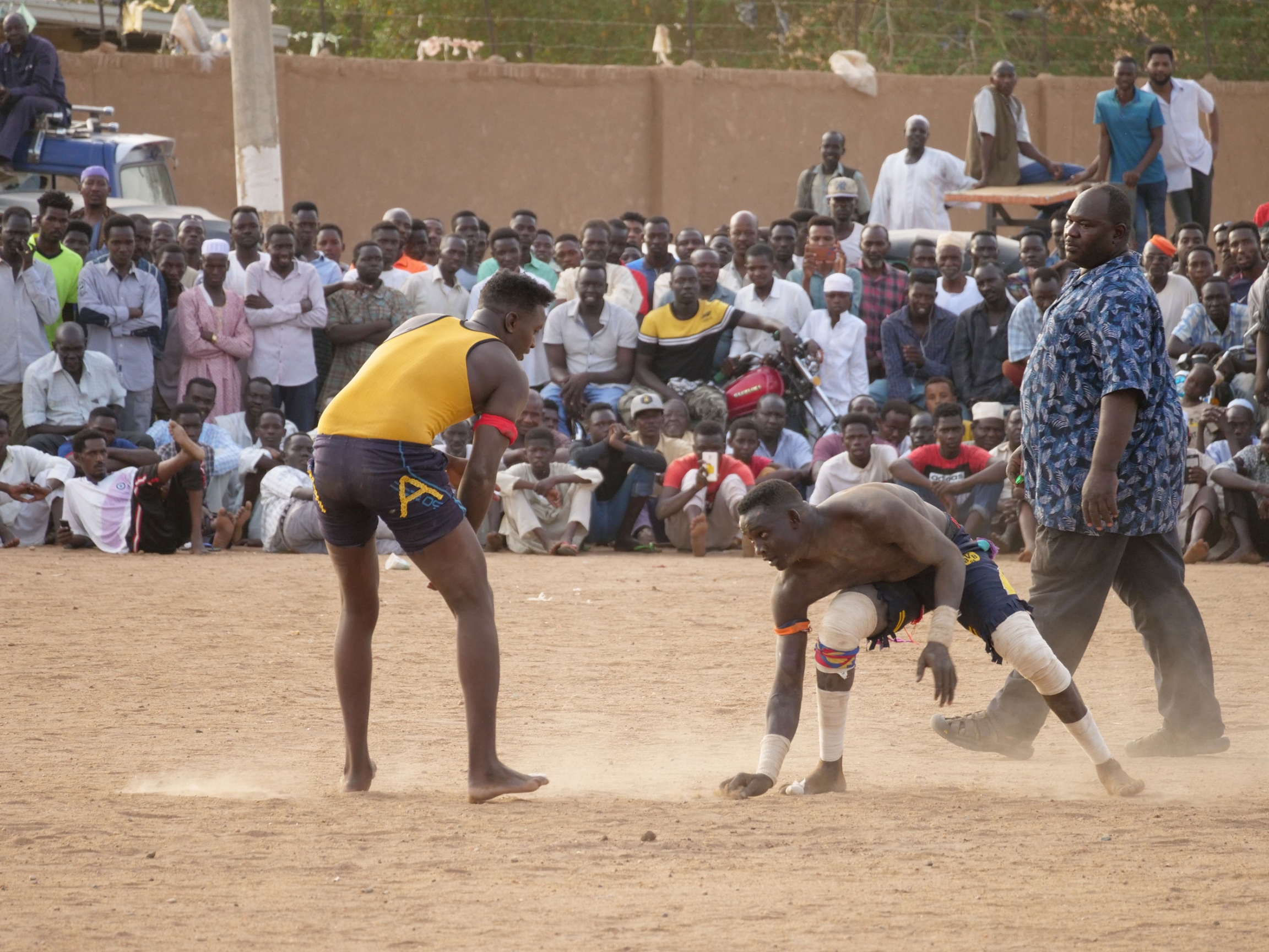 Wrestlers circle each other at a match in Khartoum, Sudan. To men are shown wearing shorts and t-shirts while wrestling in a sandy courtyard.