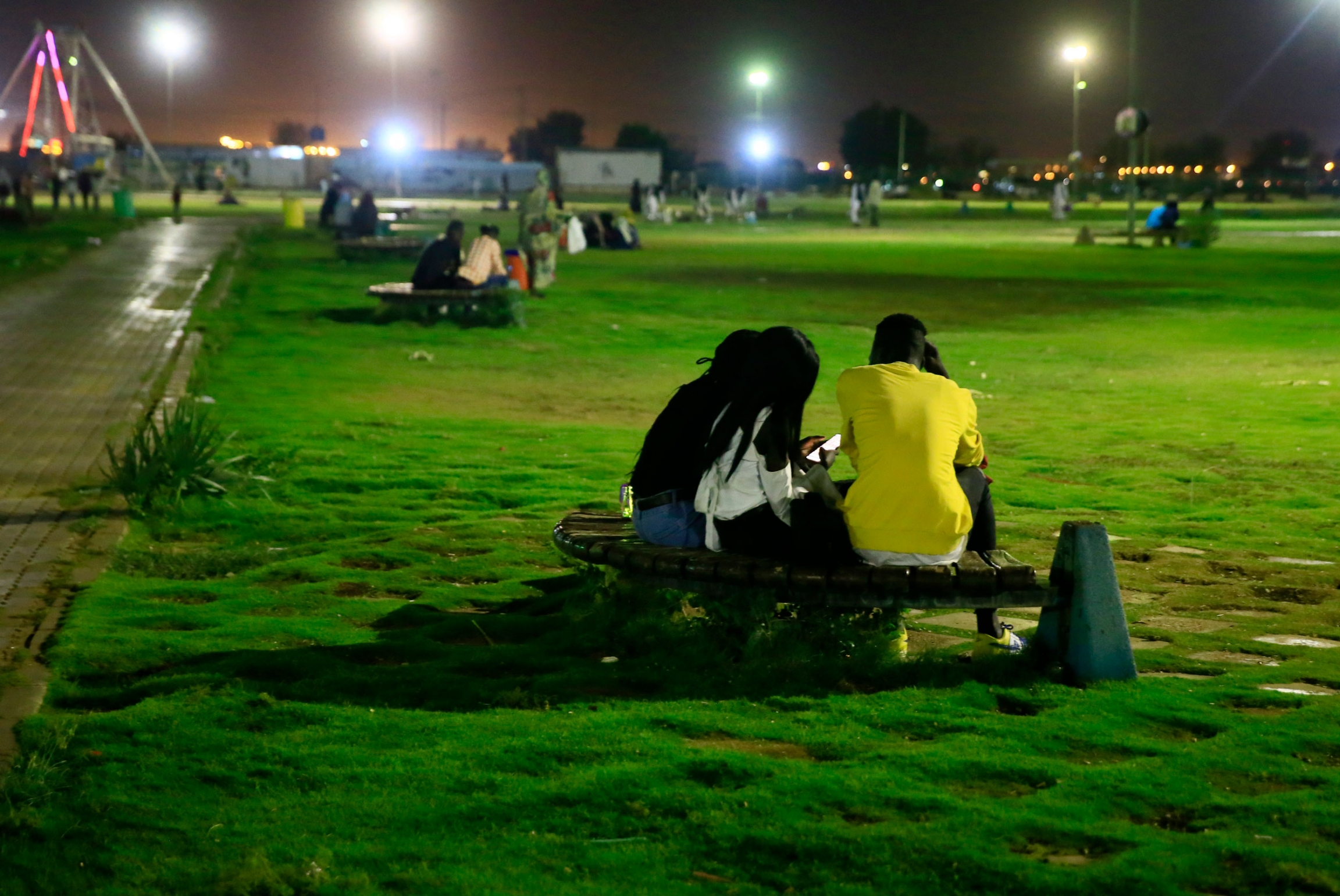 People sit on benches in Green Park at dusk in Khartoum, Sudan. Three people are shown with one person wearing a yellow shirt while sitting on a curved metal bench amid a large grassy area.