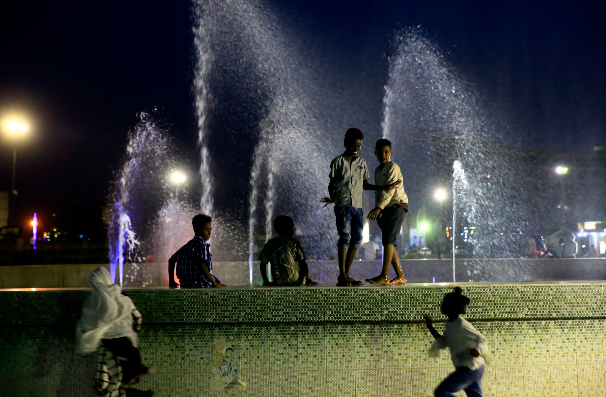 Children play near a water fountain in Khartoum, Sudan. Several children are shown standing on the tiled edge of a fountain with water spraying up in the background.