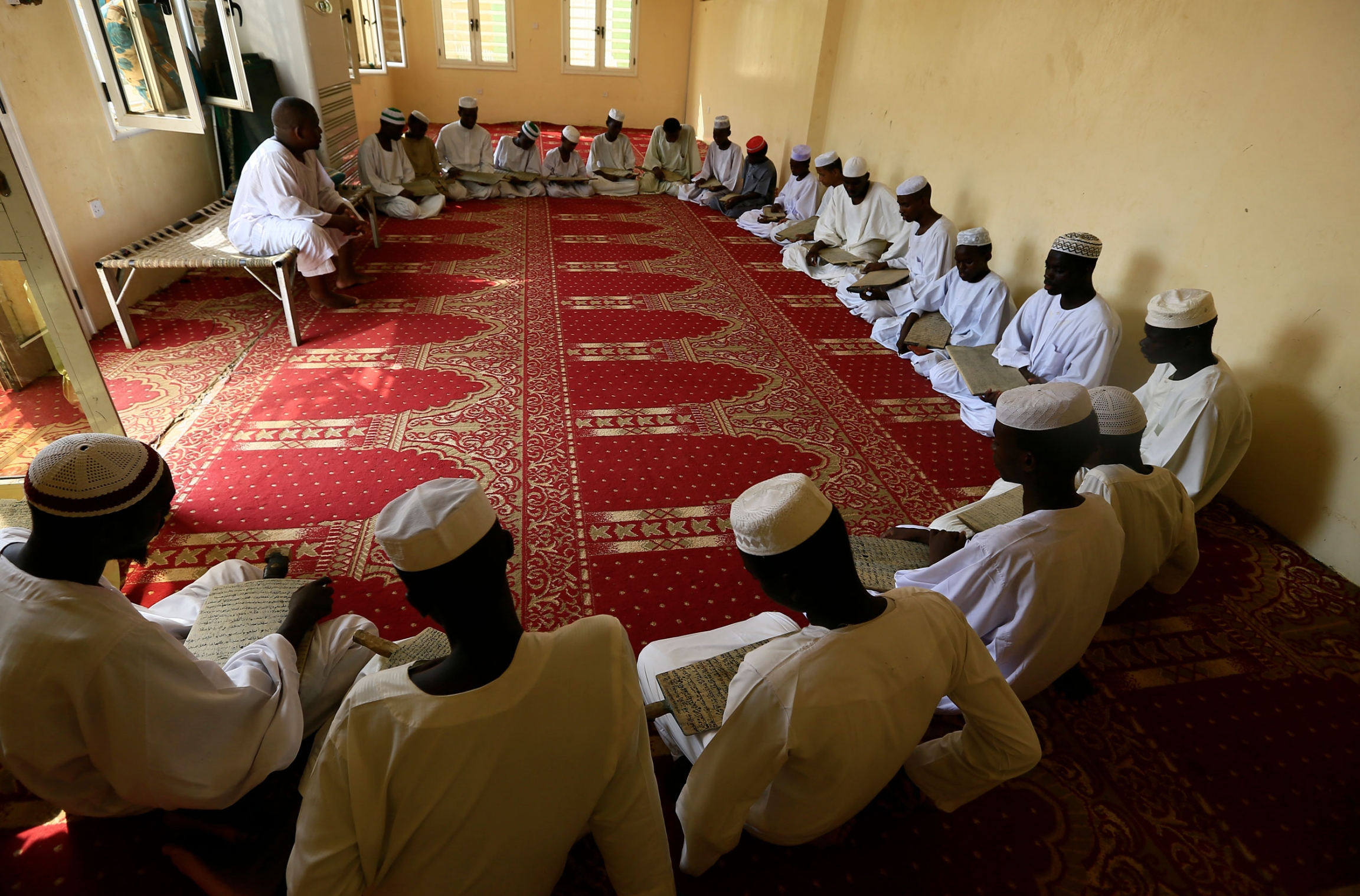 Young men sit in Haj Yousif Mosque studying Islamic text. A large group of young men are shown sitting in a half circle on the ground and holding wooden boards with Islamic text.
