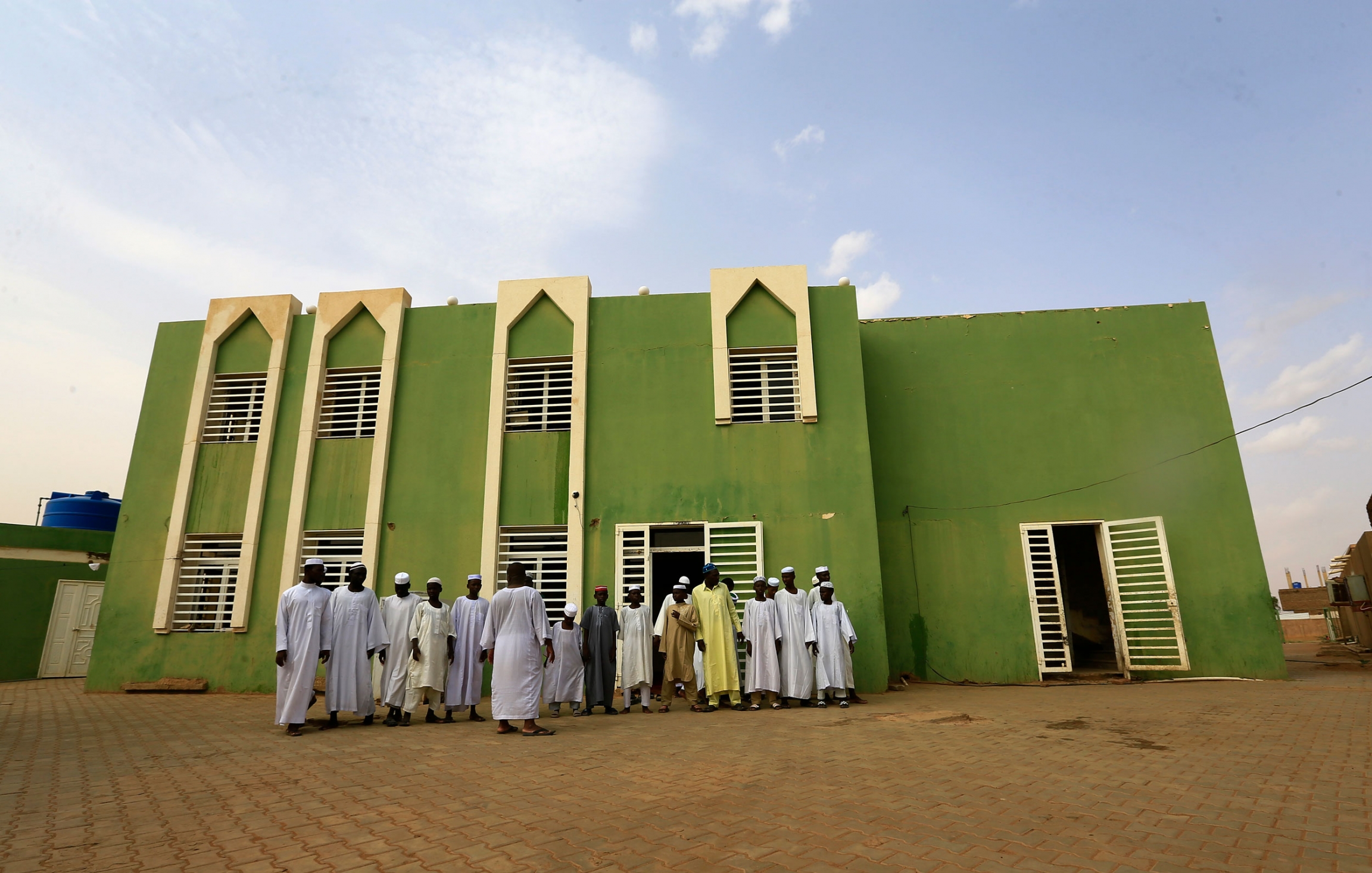 A group of young men stand outside of the Haj Yousif Mosque in Khartoum, Sudan. A group of men are shown in traditional Islamic robes outside of the green facade of the Haj Yousif Mosque.