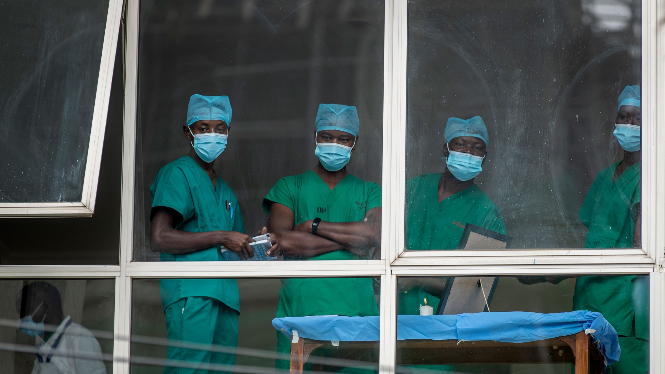Medical staff look out from a window amid the coronavirus pandemic at Kenyatta National Hospital in Nairobi, Kenya. Four people are shown looking through a white-framed windows and wearing green medical scrubs.