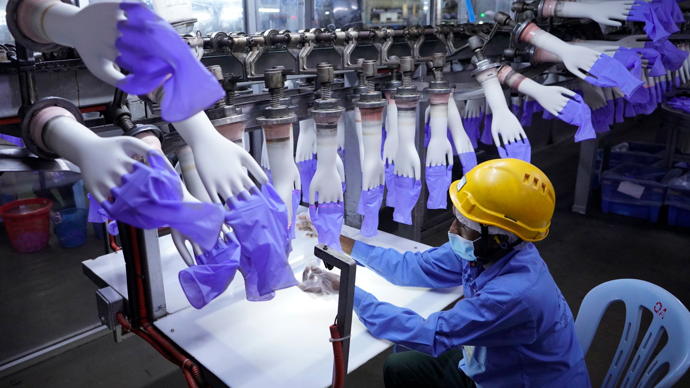 In this Aug. 26, 2020, file photo, a worker inspects disposable gloves at the Top Glove factory in Shah Alam outside Kuala Lumpur, Malaysia. A worker wearing a yellow safety hat inspects disposable blue gloves at a factory.