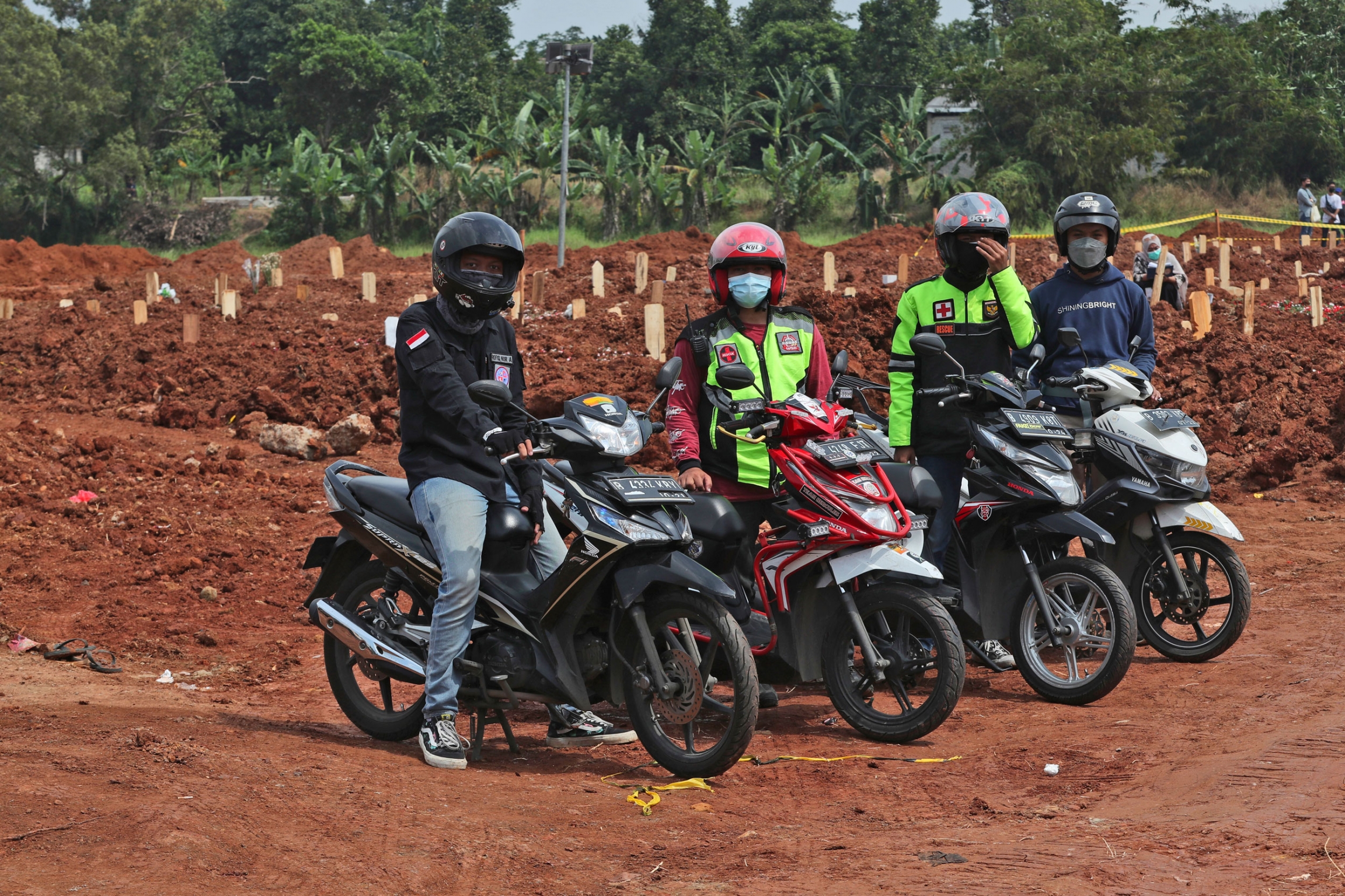 Motorcycle volunteers pose for a photo after escorting an ambulance carrying the body of a COVID-19 victim, at Pedurenan cemetery in Bekasi on the outskirts of Jakarta, Indonesia. Four people are shown sitting or standing next to motorbikes with a cemetary in the background.