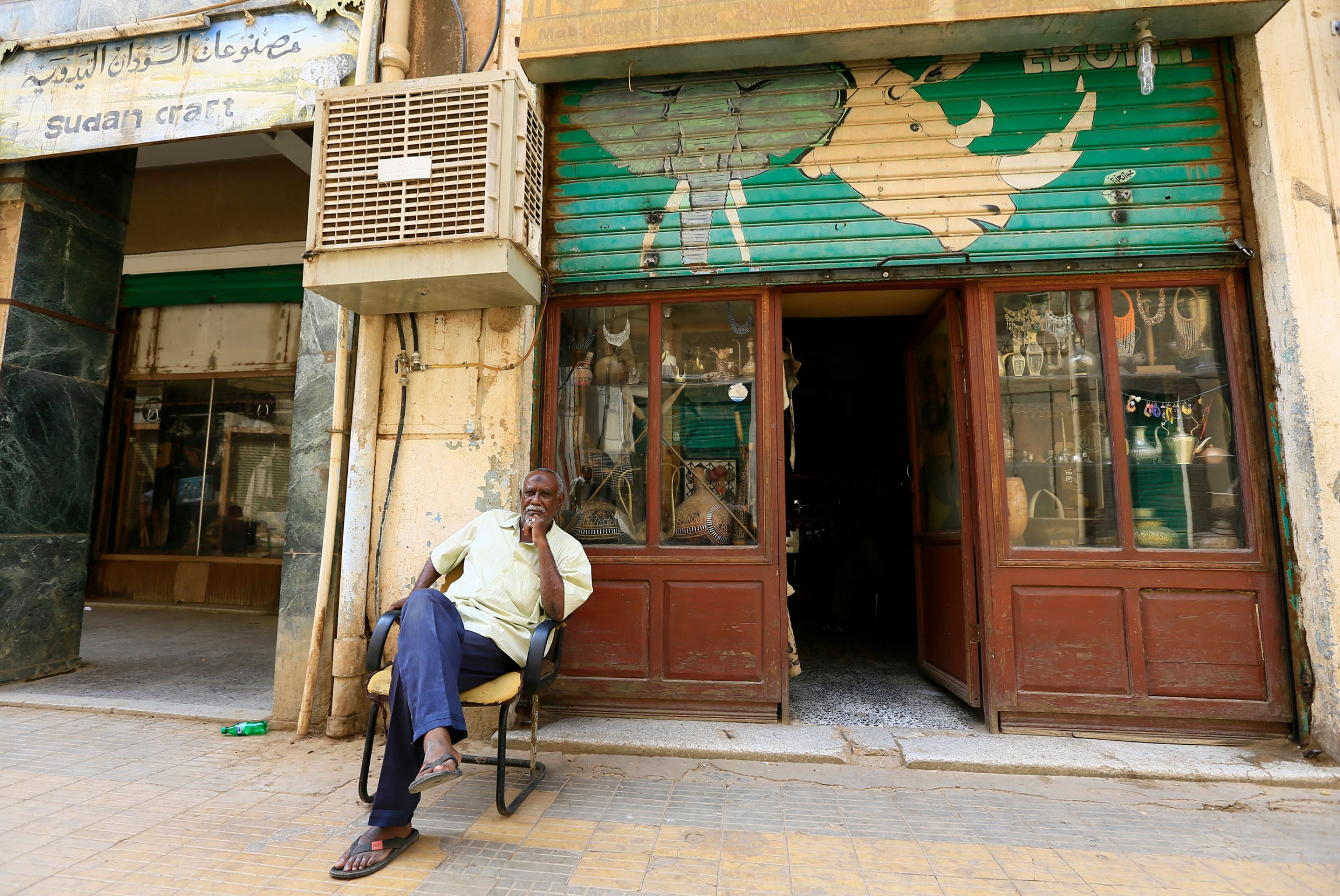 A man sits outside his antique shop in Atenay neighborhood of Khartoum, Sudan. A man is shown sitting with his legs crossed in a chair outside of a shop with windows diplaying antiques.