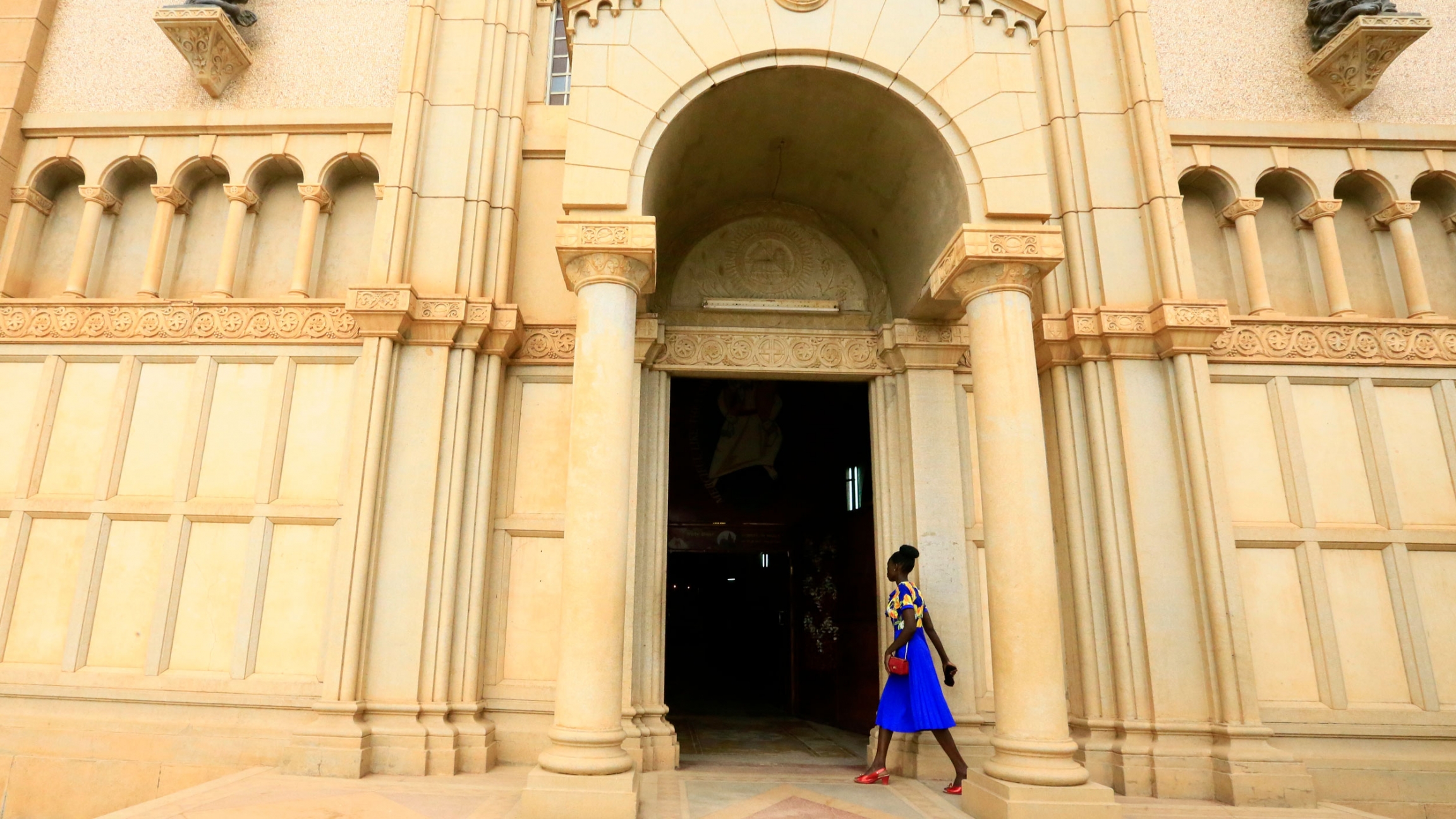 A woman walks into St. Matthew’s Catholic Cathedral in Khartoum, Sudan. A woman wearing a blue dress and red shoes walks into the tan-colored stone entrance to St. Matthew’s Catholic Cathedral