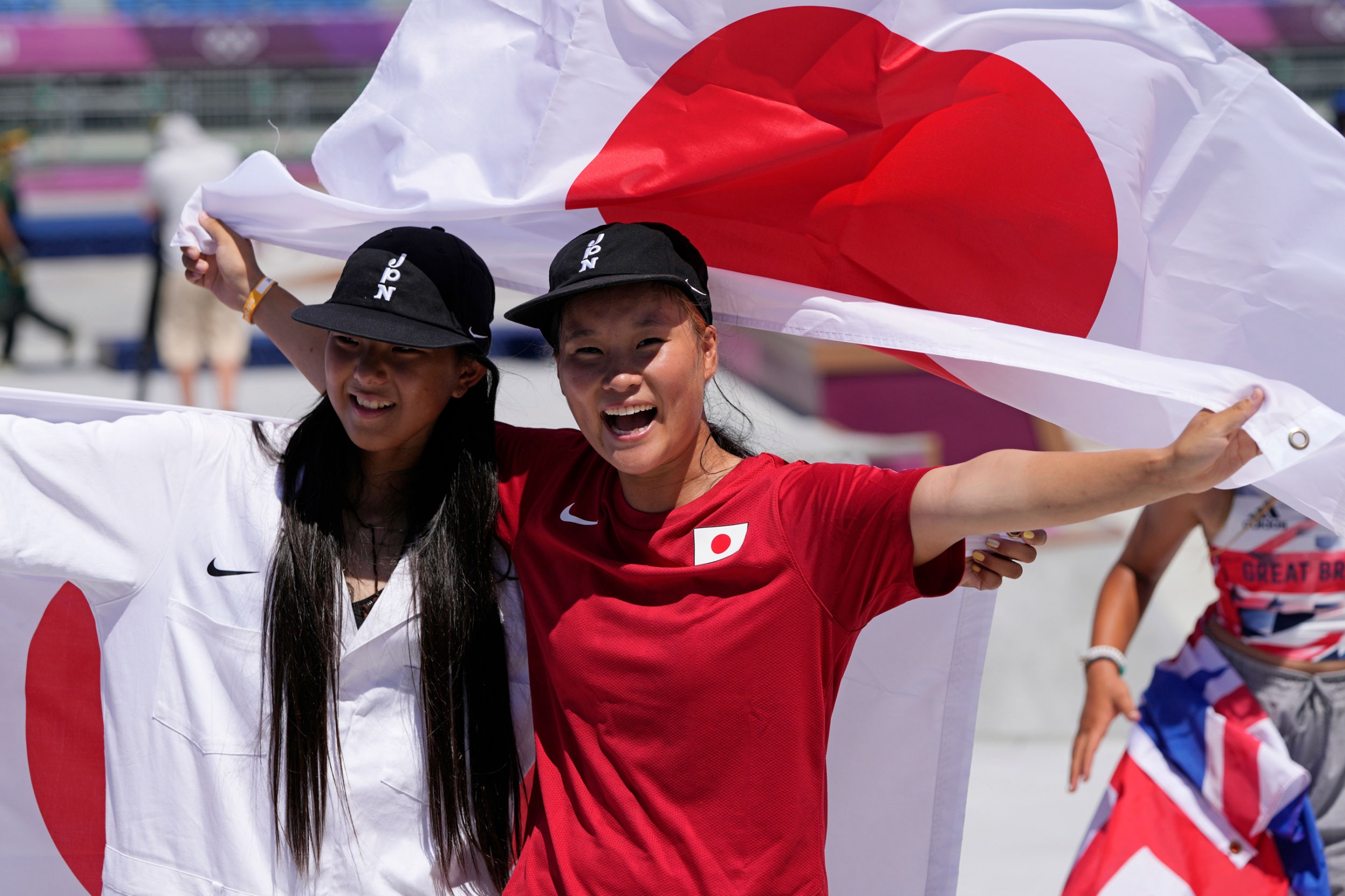 Gold medal winner Sakura Yosozumi (R) and silver medalist Kokona Hiraki, both of Japan, celebrate after the women's park skateboarding finals at the 2020 Summer Olympics, Aug. 4, 2021. Sakura Yosozumi and Kokona Hiraki are show side-by-side with a Japanese flag held up behind them.
