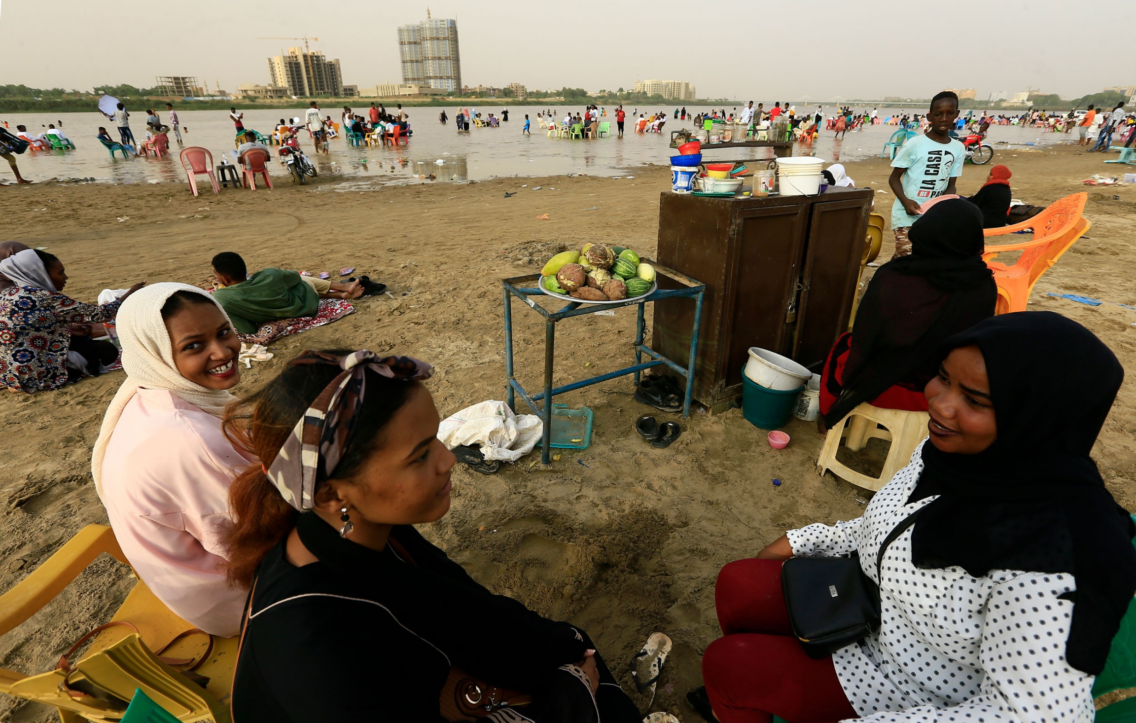A group of woman relax on the beach of the Nile River in Khartoum, Sudan. A group of three women are shown sitting with food nearby and the Nile River in the distance.