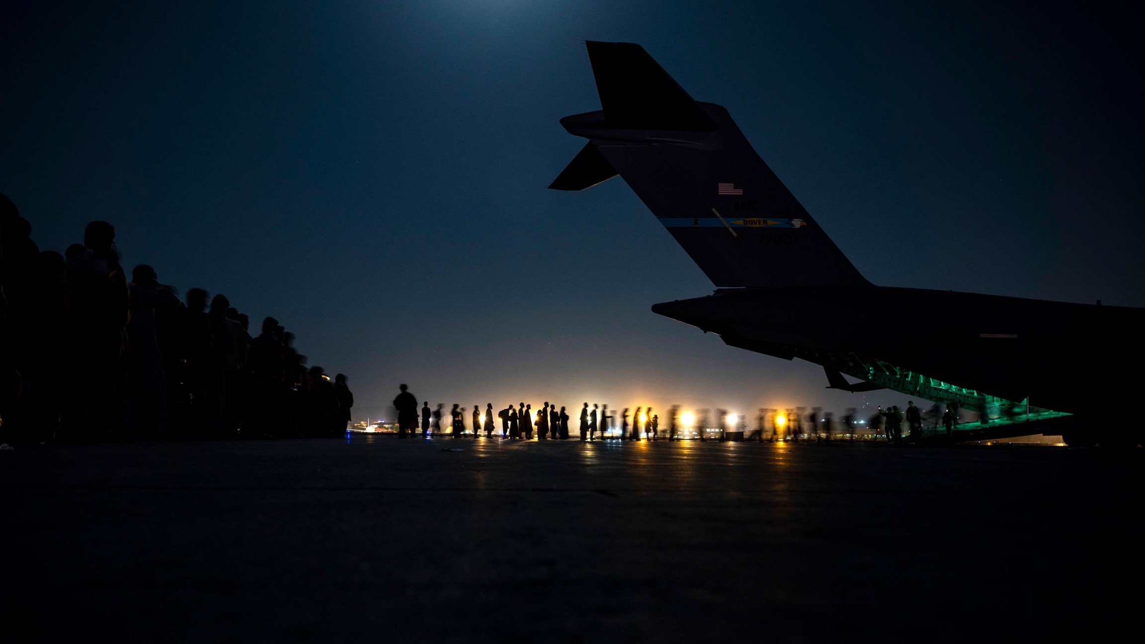 US Air Force aircrew assist evacuees boarding an Air Force C-17 aircraft at Hamid Karzai International Airport, Kabul, Afghanistan, Aug. 21, 2021. A line of people are shown in the distance in low light with the tail of a large aircraft as the sun sets.