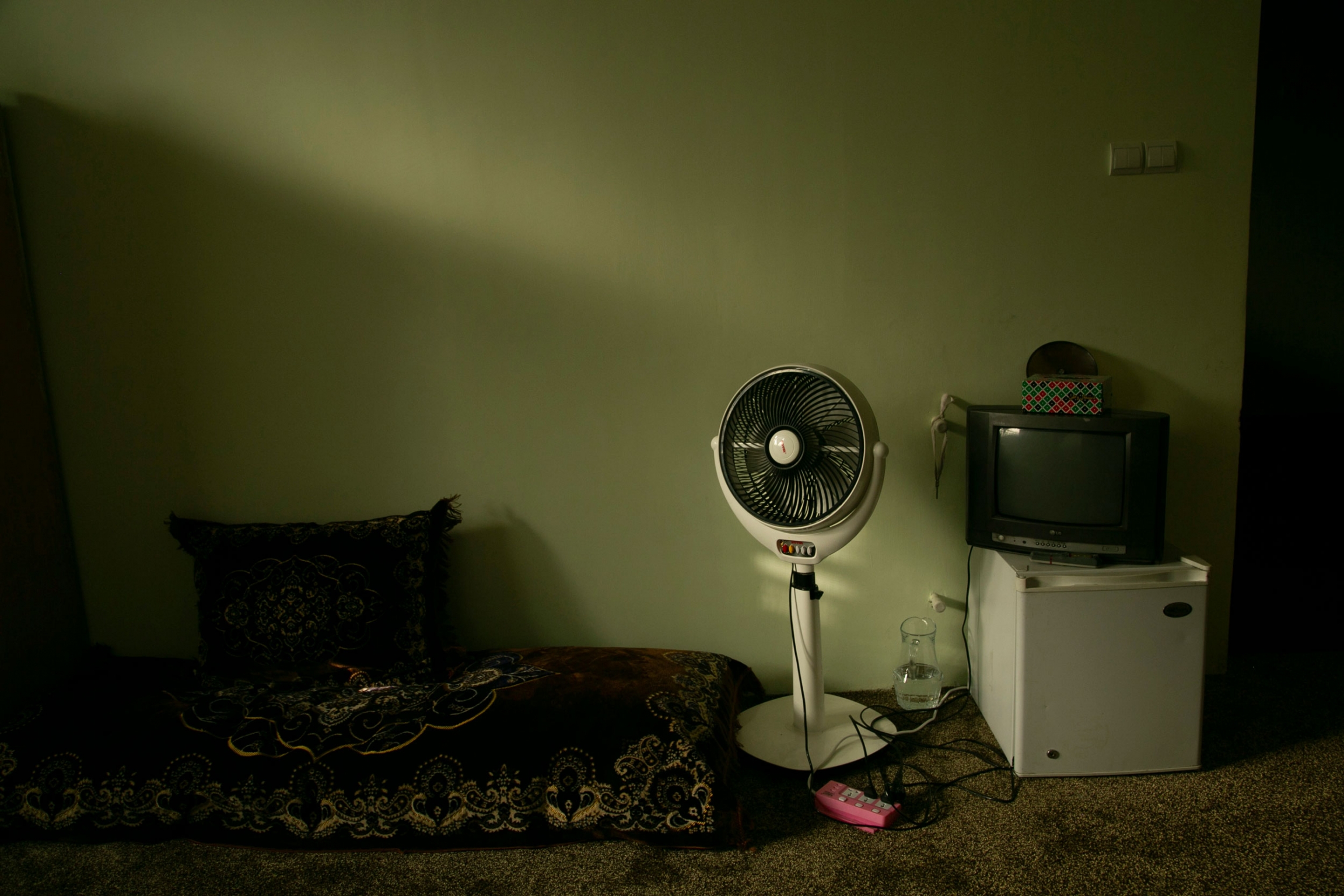 One of the rooms that women share at the shelter in Kabul, Afghanistan. A dimly lit room is shown with a large cushion and a electric fan off to the side.