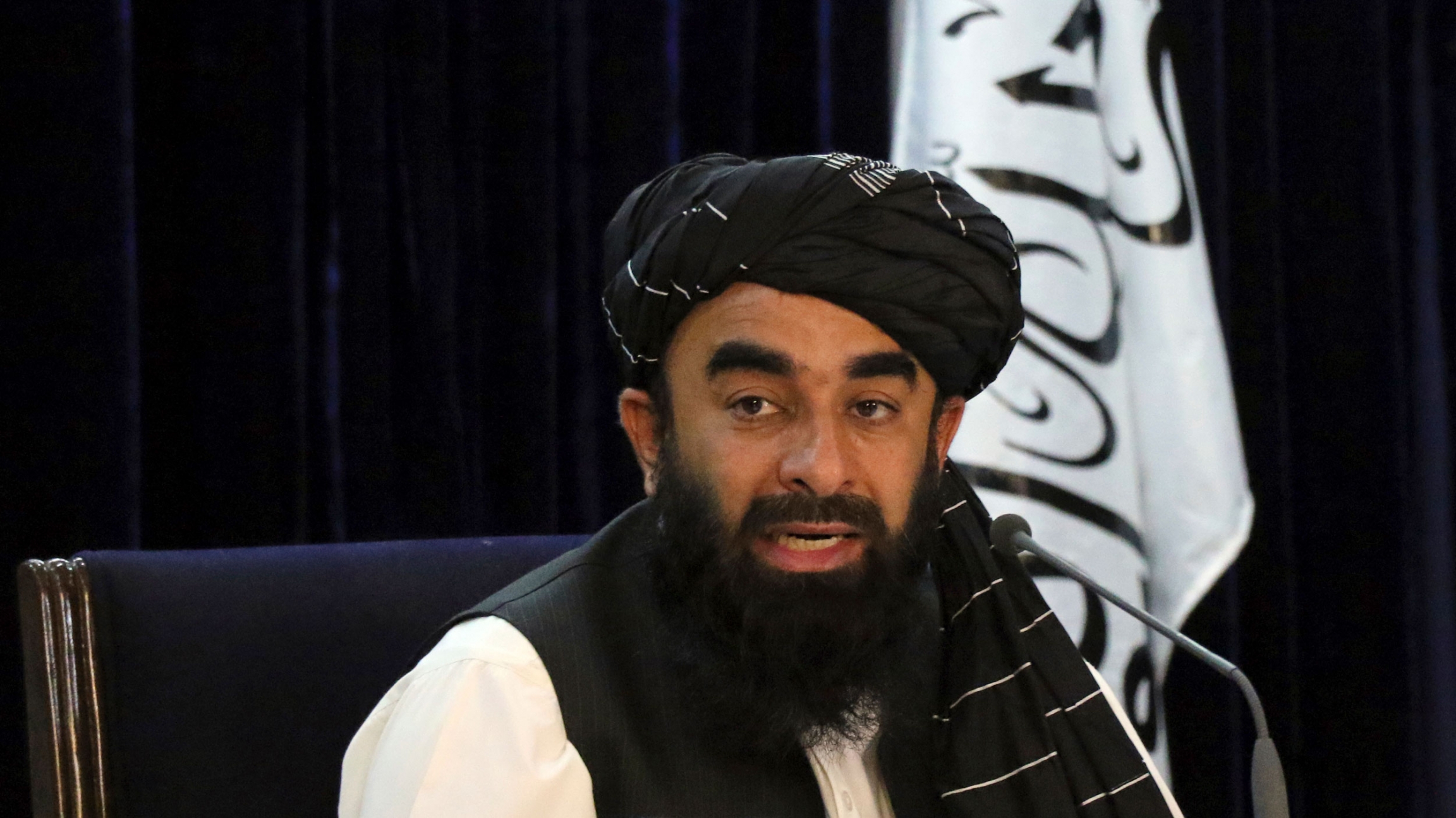 Taliban spokesman Zabihullah Mujahid speaks during a press conference in Kabul, Afghanistan, Sept. 7, 2021. The Taliban on Tuesday announced a caretaker Cabinet stacked with veterans of their harsh rule in the late 1990s. Taliban spokesman Zabihullah Mujahid is shown sitting while wearing a dark head wrap and black vest with the Taliban flag behind him.