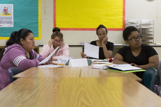 For women sit around elementary classroom tables, holding and reading papers