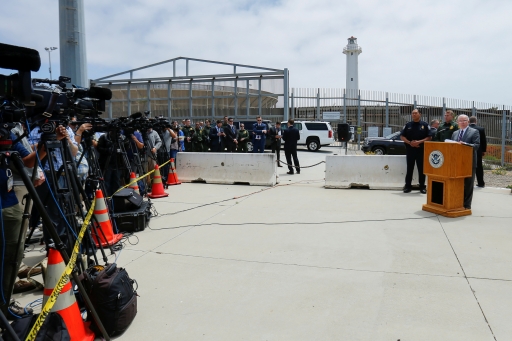 Man standing at podium, speaking, with fence behind him and people holding cameras in front
