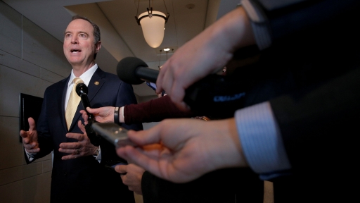 A man is lit by a bright light and surrounded by microphones as he gives an interview to a pack of reporters