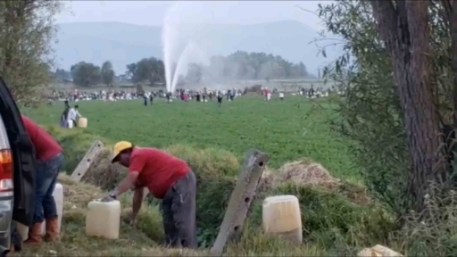 People are holding plastic gasoline containers. In the distance behind them, streams of gasoline spurts into the air out of a broken pipeline.