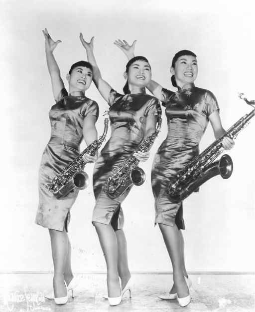 The Kim Sisters A black and white photo of three young Korean women posing with their hands up and holding saxophones.