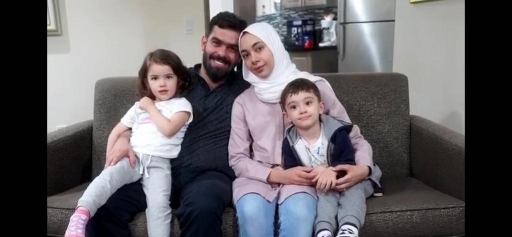 A woman wearing a hijab sits next to her husband two children on a couch.
