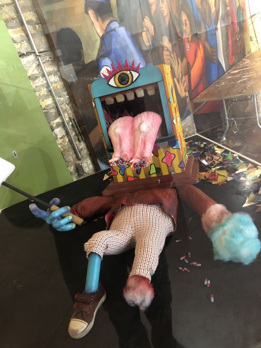 Artwork by the Puerto Rican theater collective Y No Había Luz, to be used in the haunted house. A puppet prop lays on a floor. It has a single eye, and a large pink tongue coming out of a square shaped head