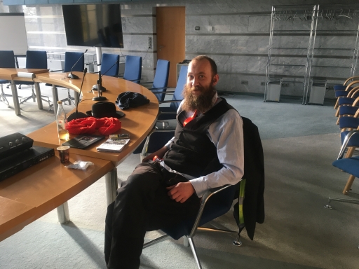 A man with a long beard and glasses in a dark suit sits at a table