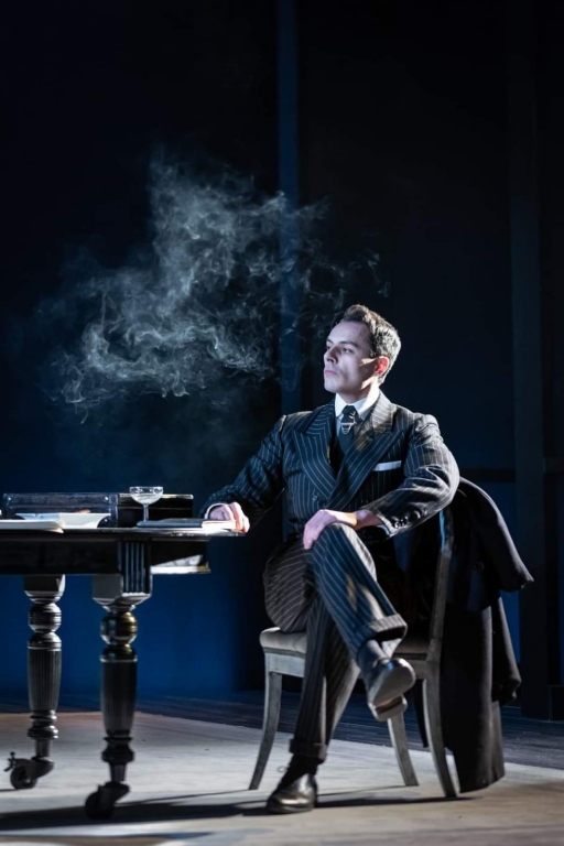 Scottish actor Noof McEwan performs in "Leopoldstadt." A man sits in a dark suit on stage with smoke around his face.