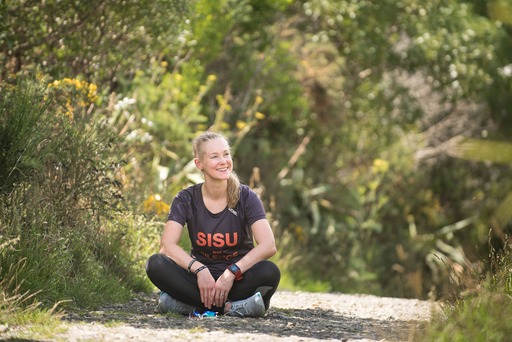 Emilia Lahti during an ultra long-distance run across the length of New Zealand in 2018. A white woman sits near lots of greenery smiling and wearing black clothes with the word sisu in red letters on her shirt.