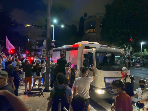 A crowd of protesters gather near a police van in Habima square in Tel Aviv. A white police van parked near a crowd of protesters.