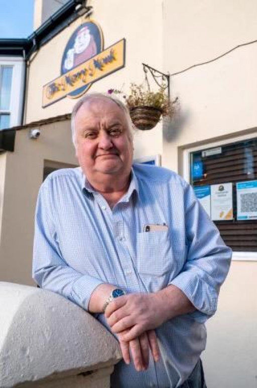 Christian Burns stands outside one of his pubs, The Merry Monk. A rotund white man with gray hair smiles and poses for a photo wearing a blue shirt outside one of his pubs, The Merry Monk.
