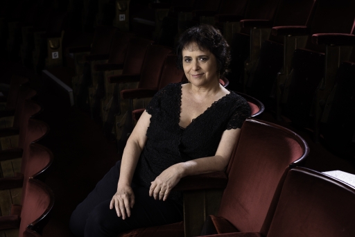 Nica Burns poses for a photo at the theater. A white woman with dark black hair wearing all black clothing poses seated in a chair at a theater, smiling to the camera with her arms on her lap.