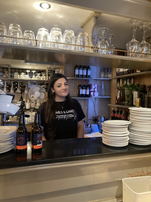 Giada Calabrese, from southern Italy, arrived in London in February 2020. She says the past year has been tough, but that she feels lucky to have a job. Giada Calabrese, from southern Italy, works behind the bar in a black shirt surrounded by cups and plates.