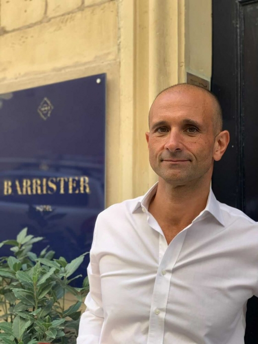 Mark Gauci is the owner of The Barrister, a boutique hotel in Valletta, Malta. Mark Gauci wears a white button-down shirt near a blue sign that reads "The Barrister," his hotel.
