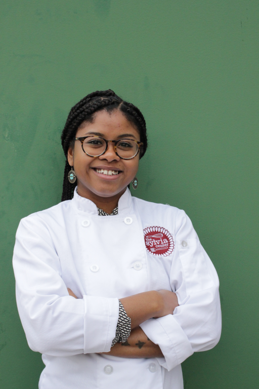 Woman against green backdrop standing and smiling, arms folded, wearing a chef's jacket